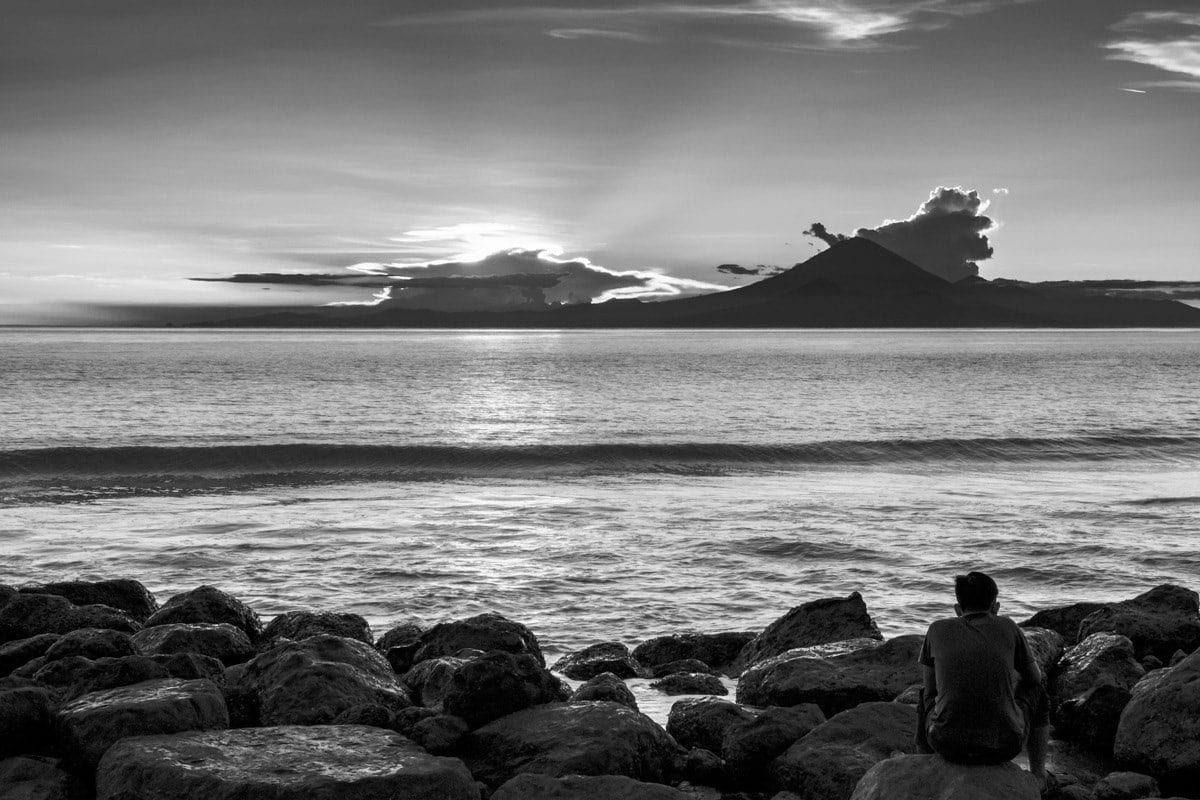Man seats on rocks and looks at Mount Agung at sunset. Lombok Island, Indonesia.