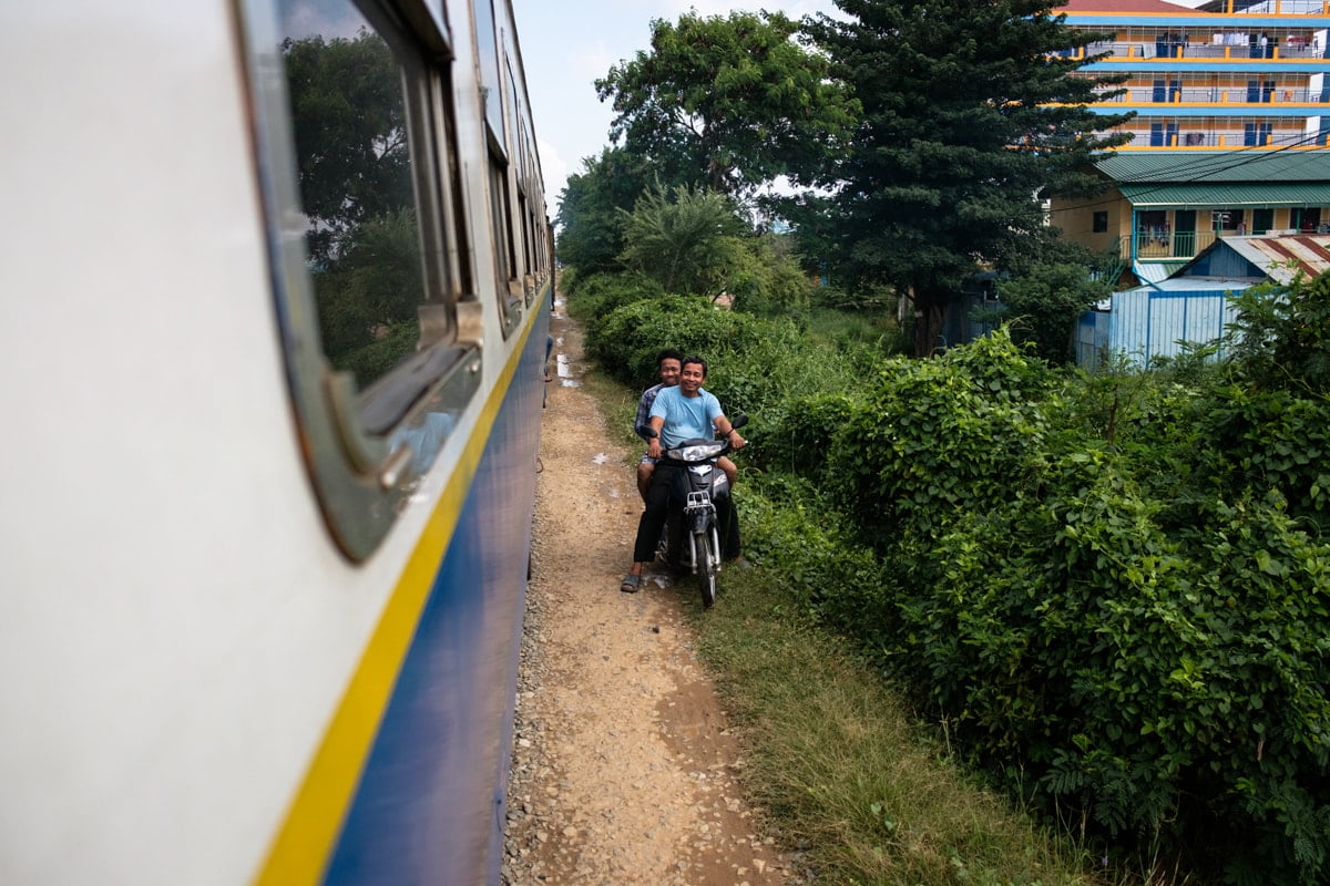 Two Cambodian men driving on a scooter next to the railway right along the train smile for my camera. Cambodia.
