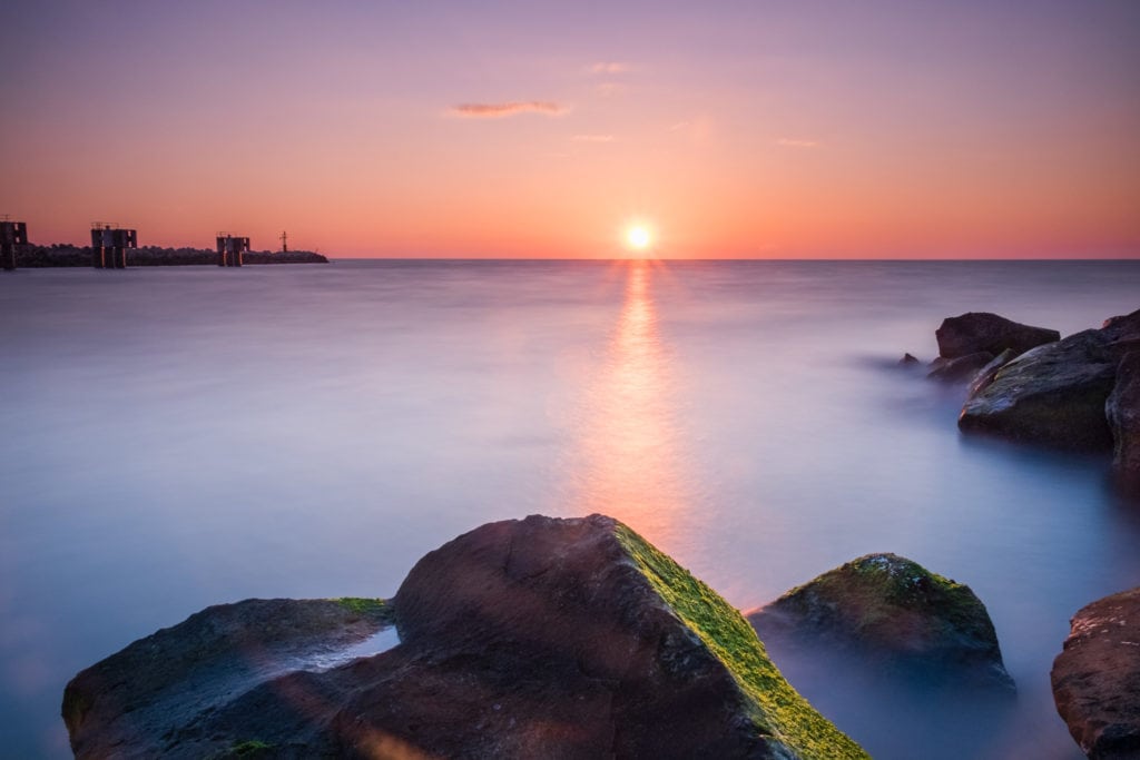 Long exposure of Rocks in the sea at sunset at Molo di Fiumicino. The sun is setting behind the sea horizon at Fiumicino Pier. With a long exposure the sea water looks smooth and silky. Rome, Italy.