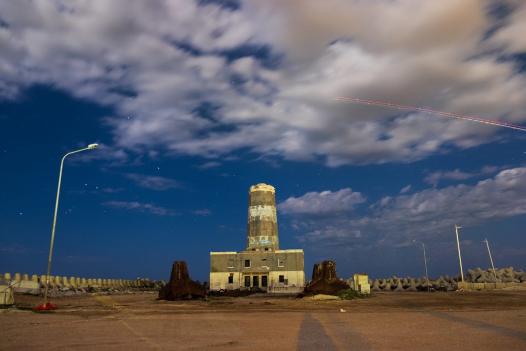 Old lighthouse of Fiumicino by night (Time 21:24). Rome, Italy. The lighthouse is lit by the moonlight and an aeroplane left light trails through the sky.